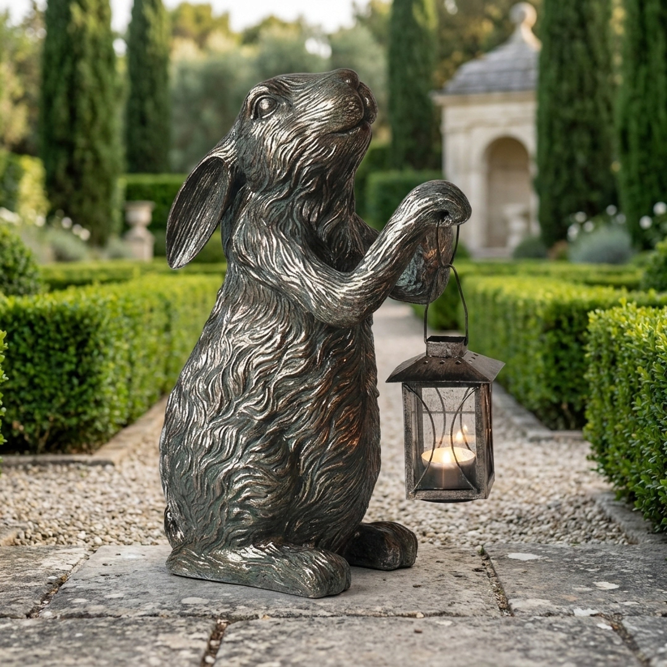 Traditional Rabbit with Lantern Bronze Sculpture by Giant Sculptures, 50cm, shown on a gravel garden path lined with hedges and cypress trees.