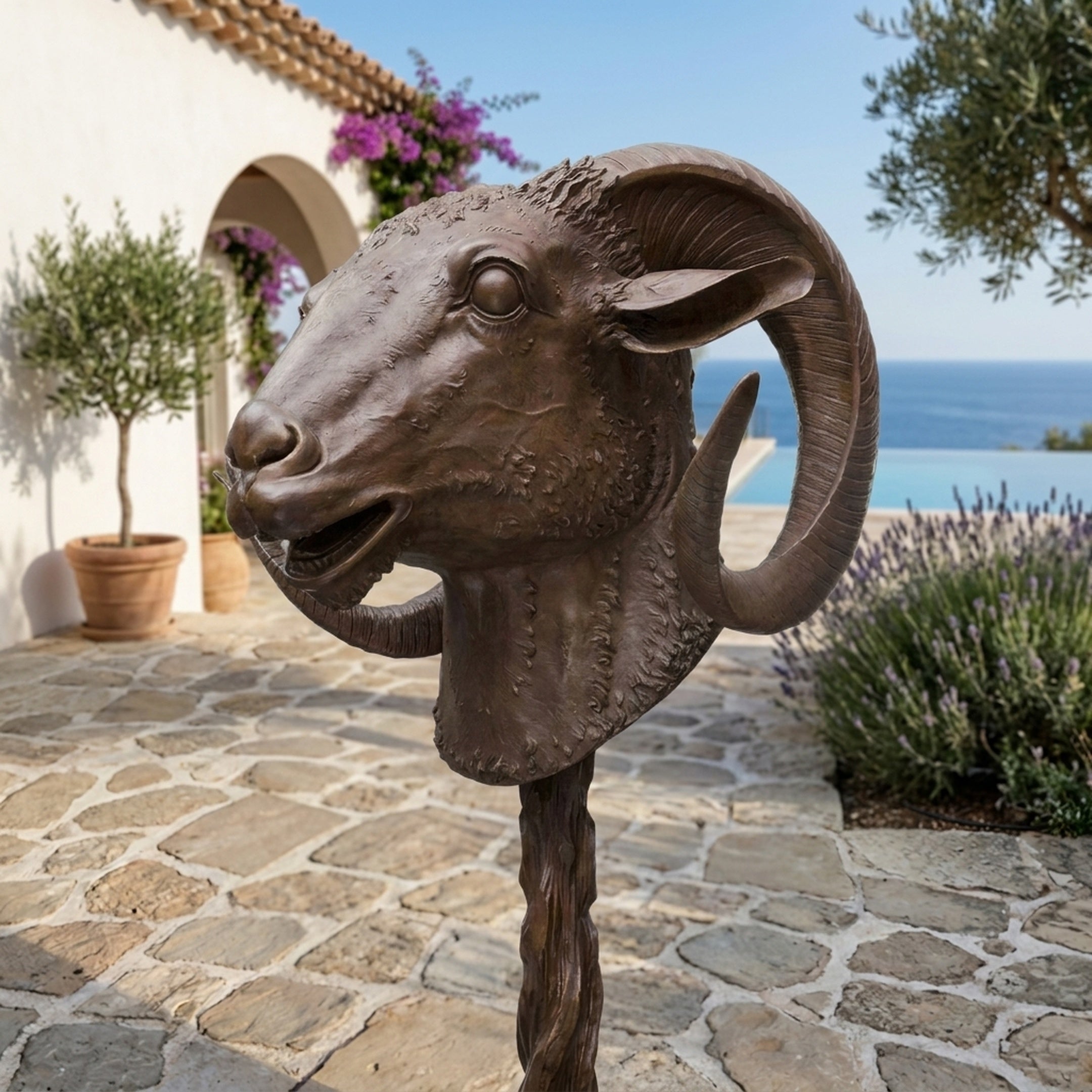 Traditional Ram Head Bronze Sculpture by Giant Sculptures shown in profile on a twisted shaft, displayed on a stone terrace with sea views.