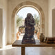 Traditional Reading Monkey Bronze Sculpture by Giant Sculptures displayed on a rustic wooden table beside leather books, with a stone archway opening to an olive grove behind.