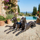Traditional Reclining Greyhound Pair Bronze Sculpture by Giant Sculptures, shown on a stone terrace beside potted flowers and an infinity pool overlooking the sea.
