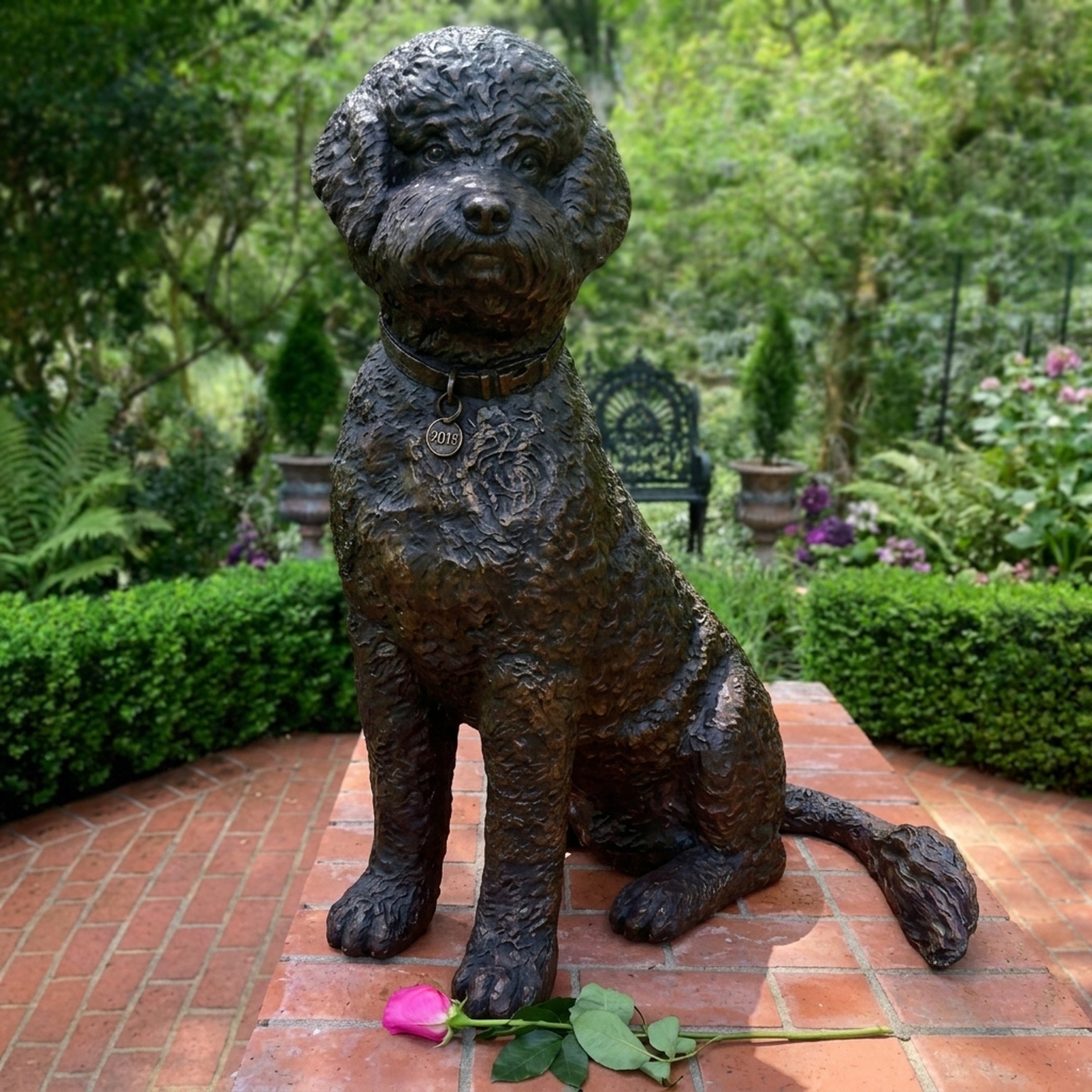 Traditional Seated Curly-Haired Dog Bronze Sculpture by Giant Sculptures on a brick terrace, with a pink rose laid at its paws and a garden behind.