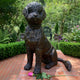 Traditional Seated Curly-Haired Dog Bronze Sculpture by Giant Sculptures on a brick terrace, with a pink rose laid at its paws and a garden behind.