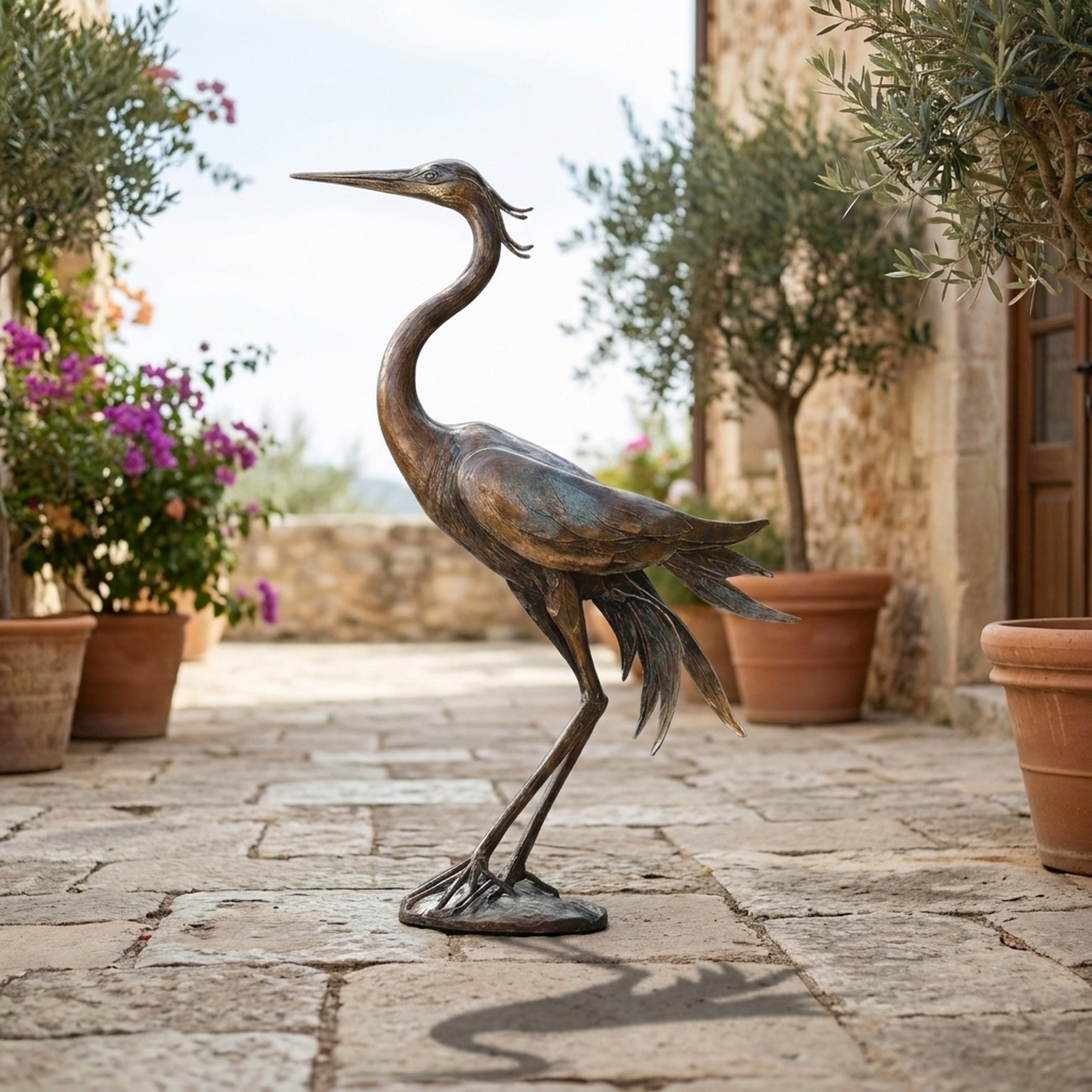 Traditional Standing Heron Bronze Sculpture by Giant Sculptures, 110cm tall with verdigris patina, displayed on a stone terrace beside potted olive trees and bougainvillea.