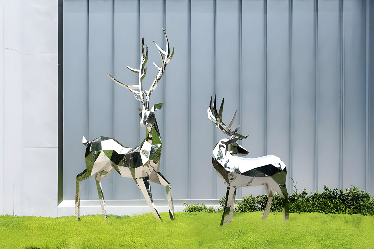 Two metallic deer sculptures with a geometric design stand on a grassy surface in front of a corrugated metal wall. One deer has larger antlers and appears taller, while the other is slightly smaller and lower to the ground.